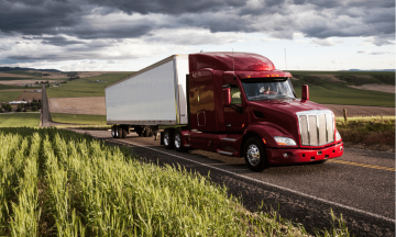 Red semi-truck transporting goods on rural road, surrounded by green fields and cloudy sky, representing domestic transportation services by Smith Transport Solutions.