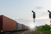 Cargo train with shipping containers on tracks, signaling lights in foreground, representing efficient rail freight solutions for logistics and supply chain management.